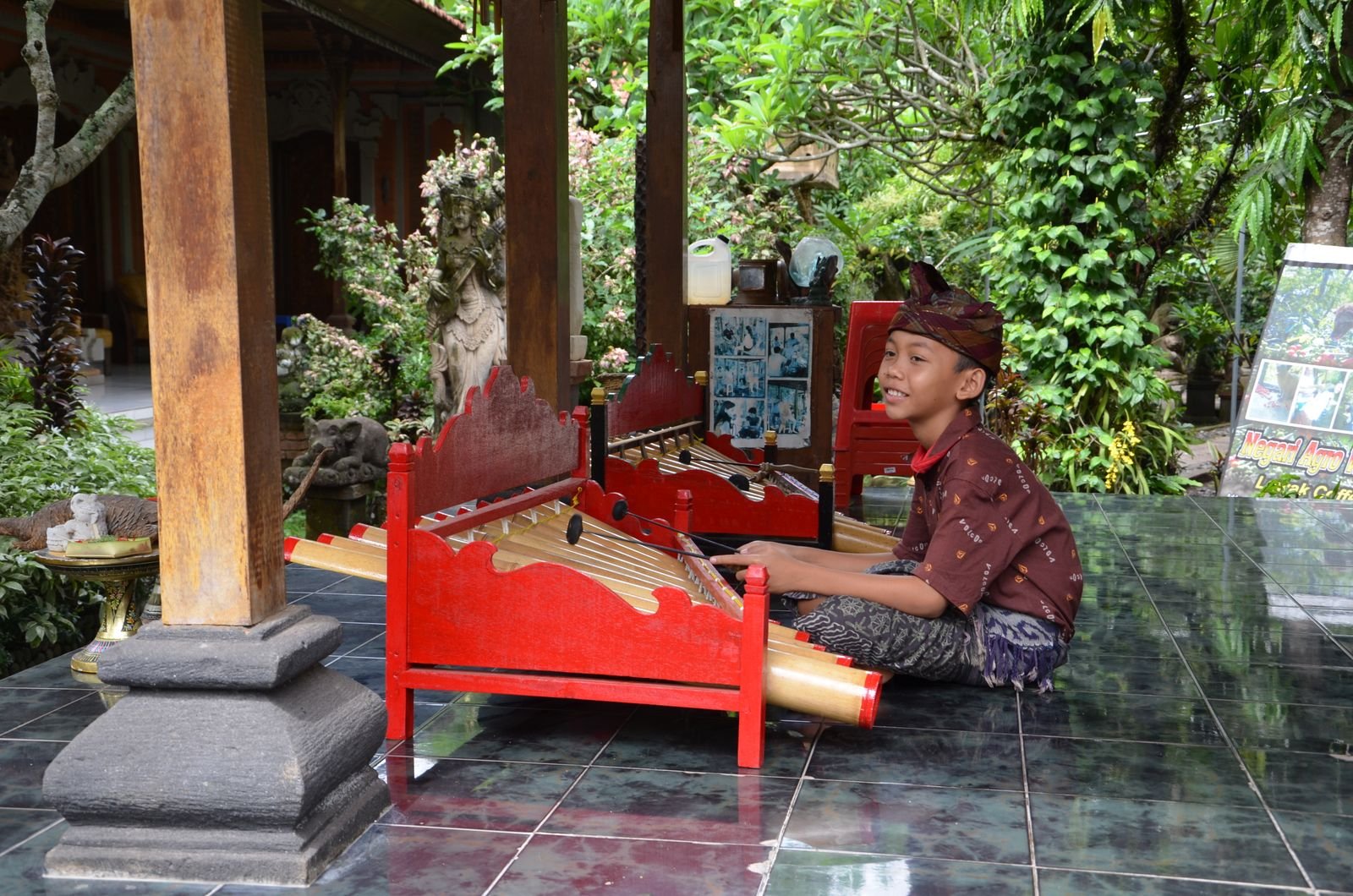 A Balinese boy in a batik shirt and udeng playing a red-framed gamelan instrument in an open pavilion
