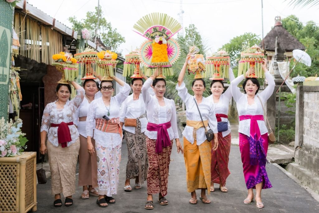 Balinese women in white kebaya and bright sashes carrying tall fruit and flower offerings on their heads along a village lane