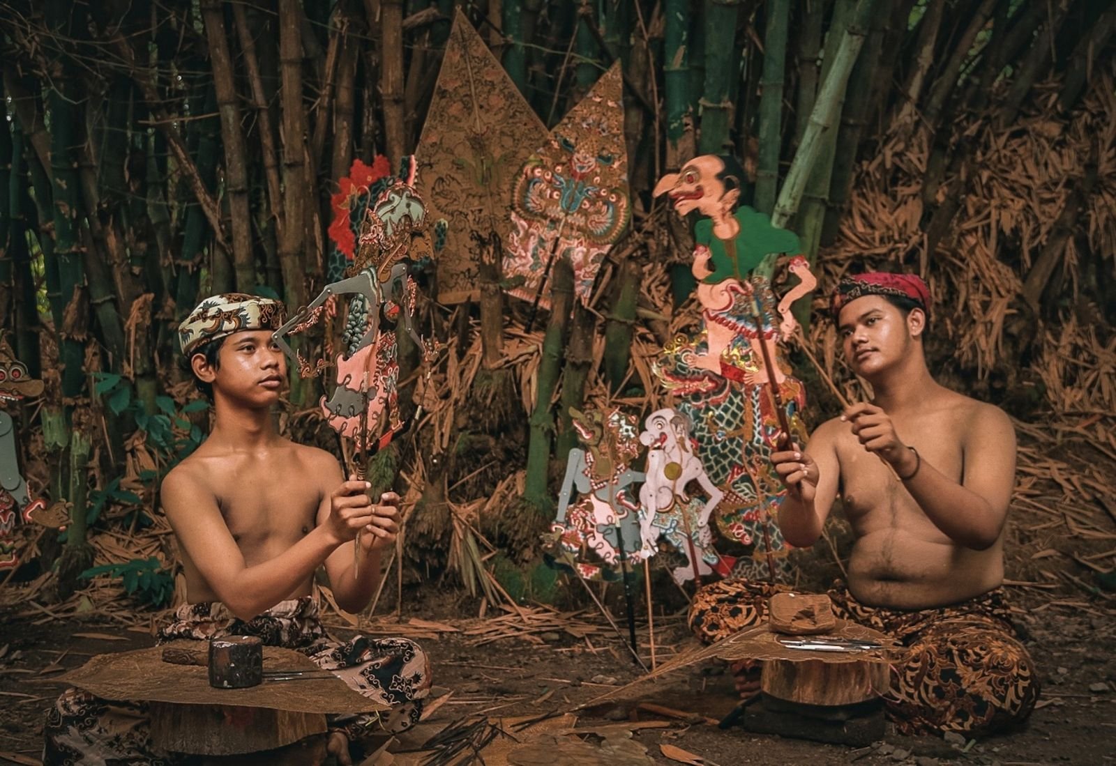 Two young performers holding painted wayang kulit shadow puppets in front of a bamboo backdrop